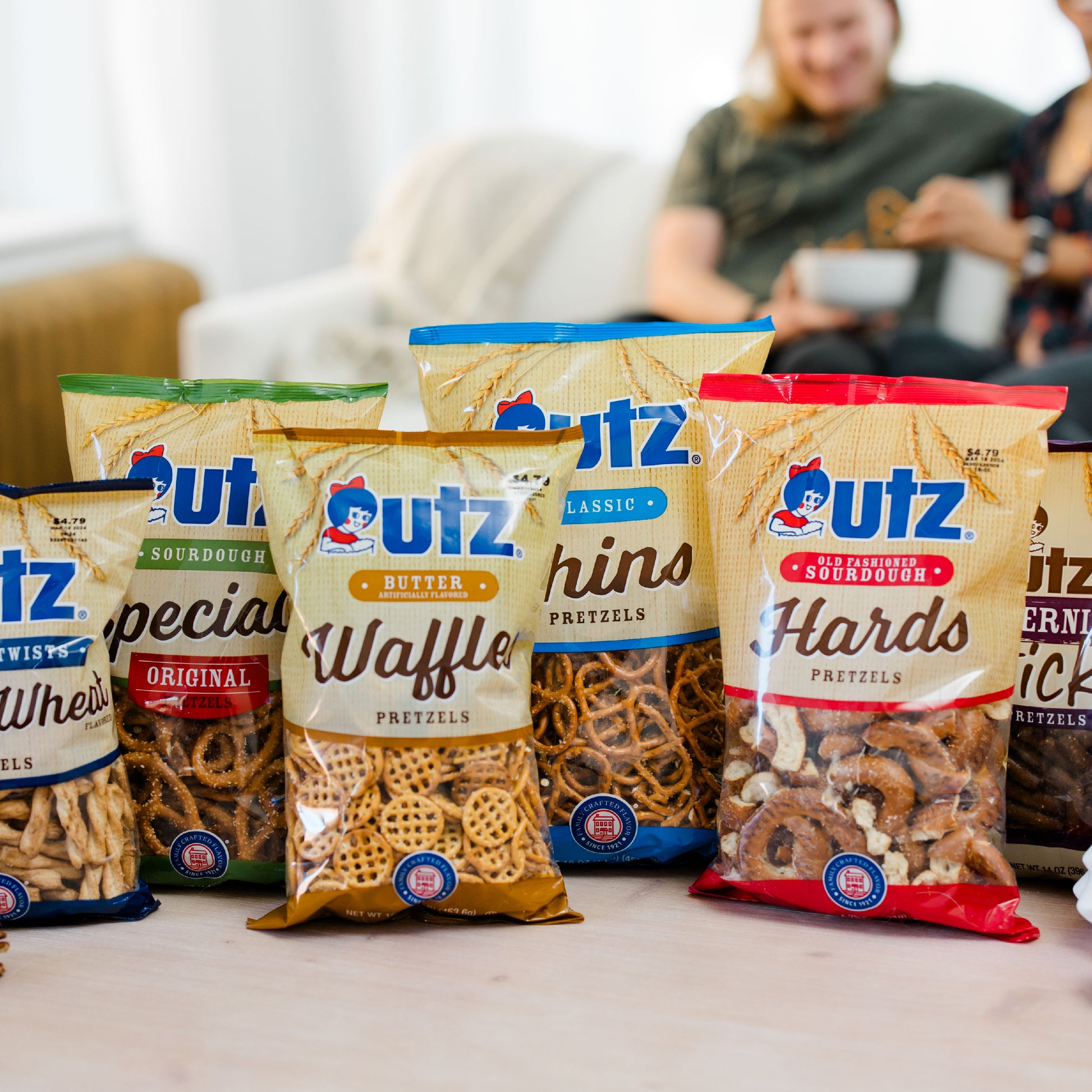 Assorted Utz pretzel packages on a table with a blurred background of people sitting on a couch.