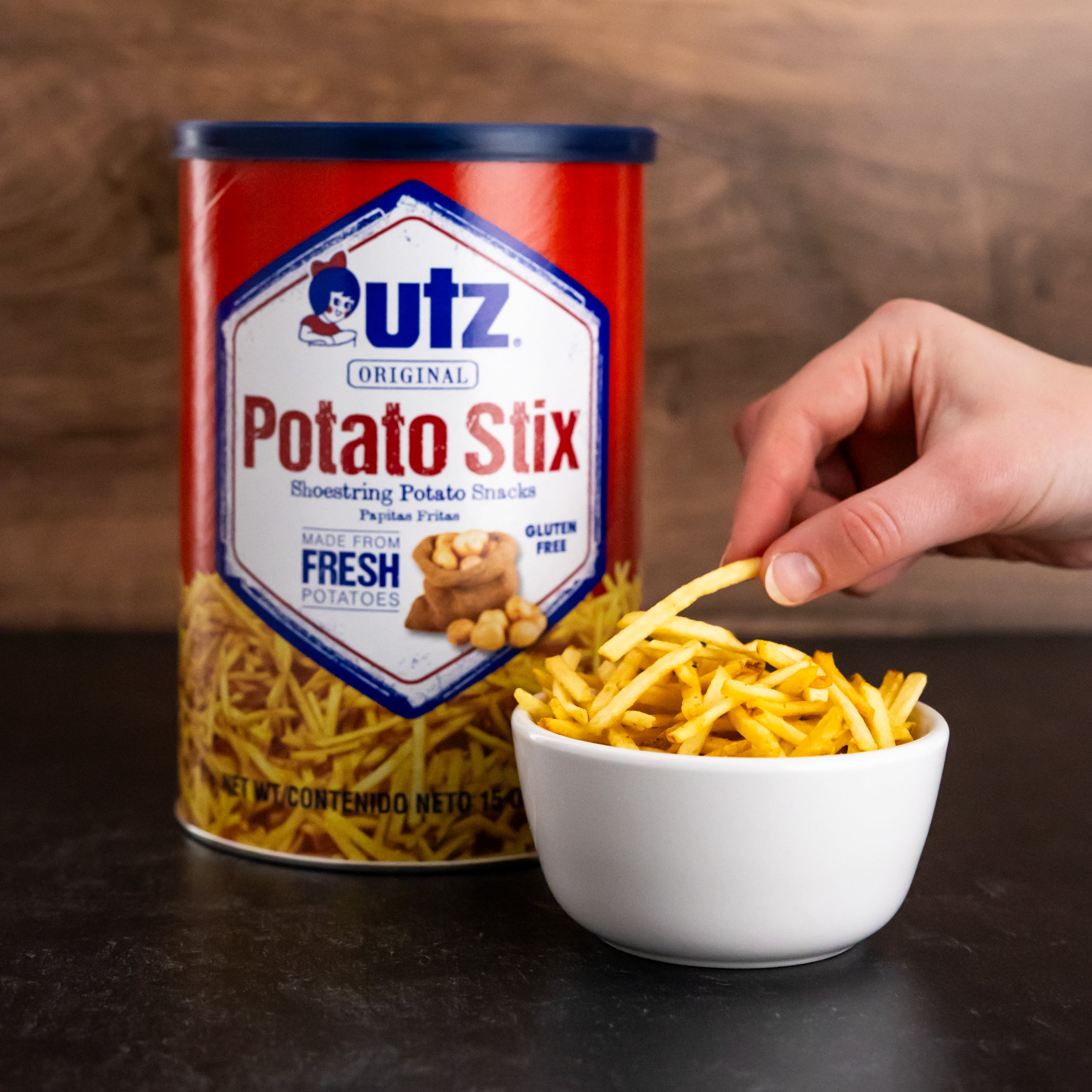 Image of a 15 oz canister of Utz Original Potato Stix beside a small white bowl filled with potato sticks. A hand is reaching into the bowl to grab some potato sticks. The canister label reads 'Made from Fresh Potatoes' and 'Gluten Free.'