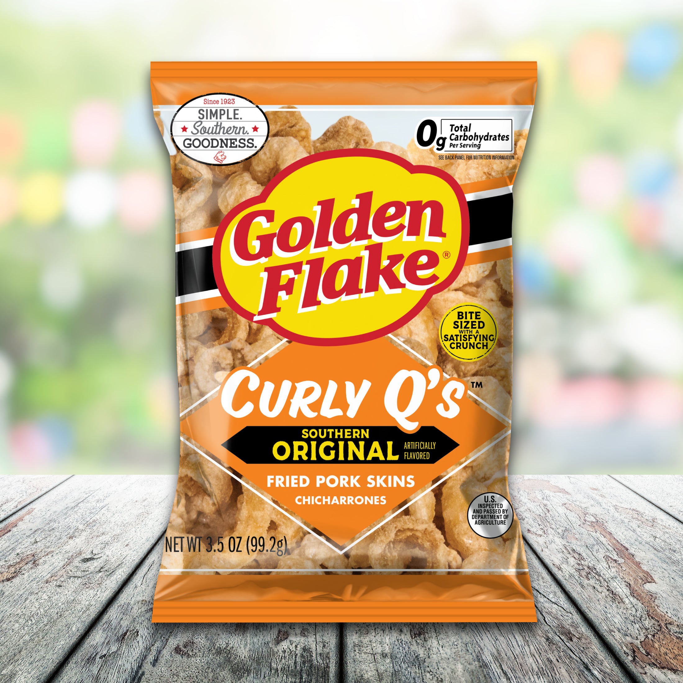 Packaging of Golden Flake Curly Q's fried pork skins on a wooden surface with a blurred colorful background.