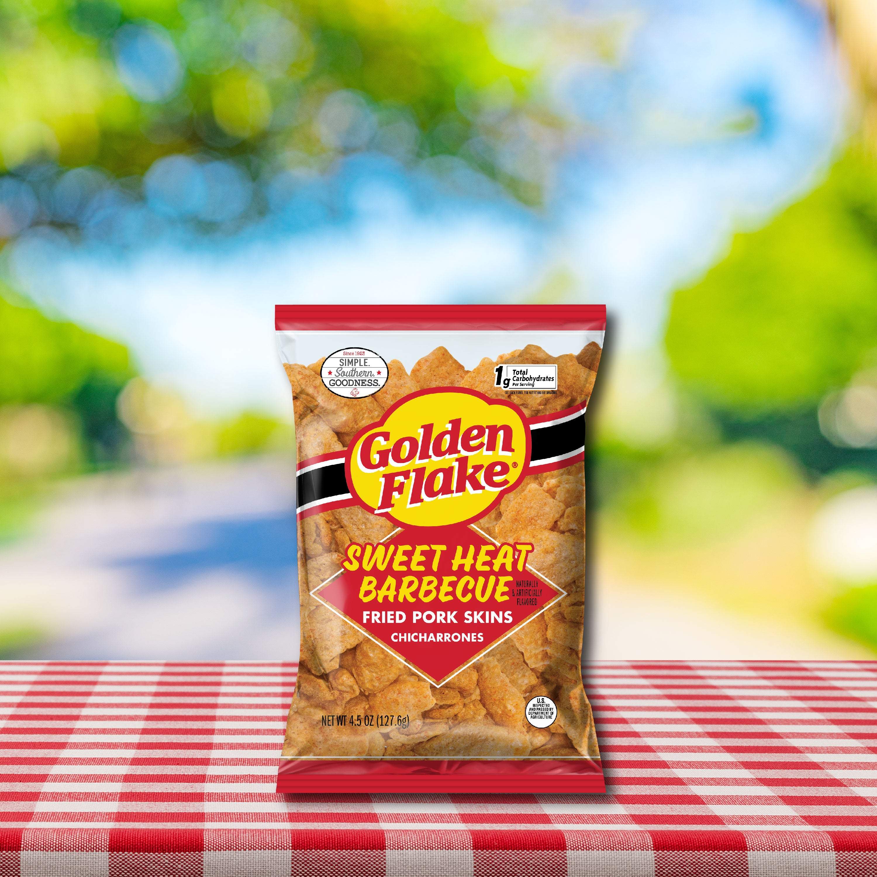 Bag of Golden Flake Sweet Heat Barbecue fried pork skins on a checkered tablecloth with a blurred outdoor background