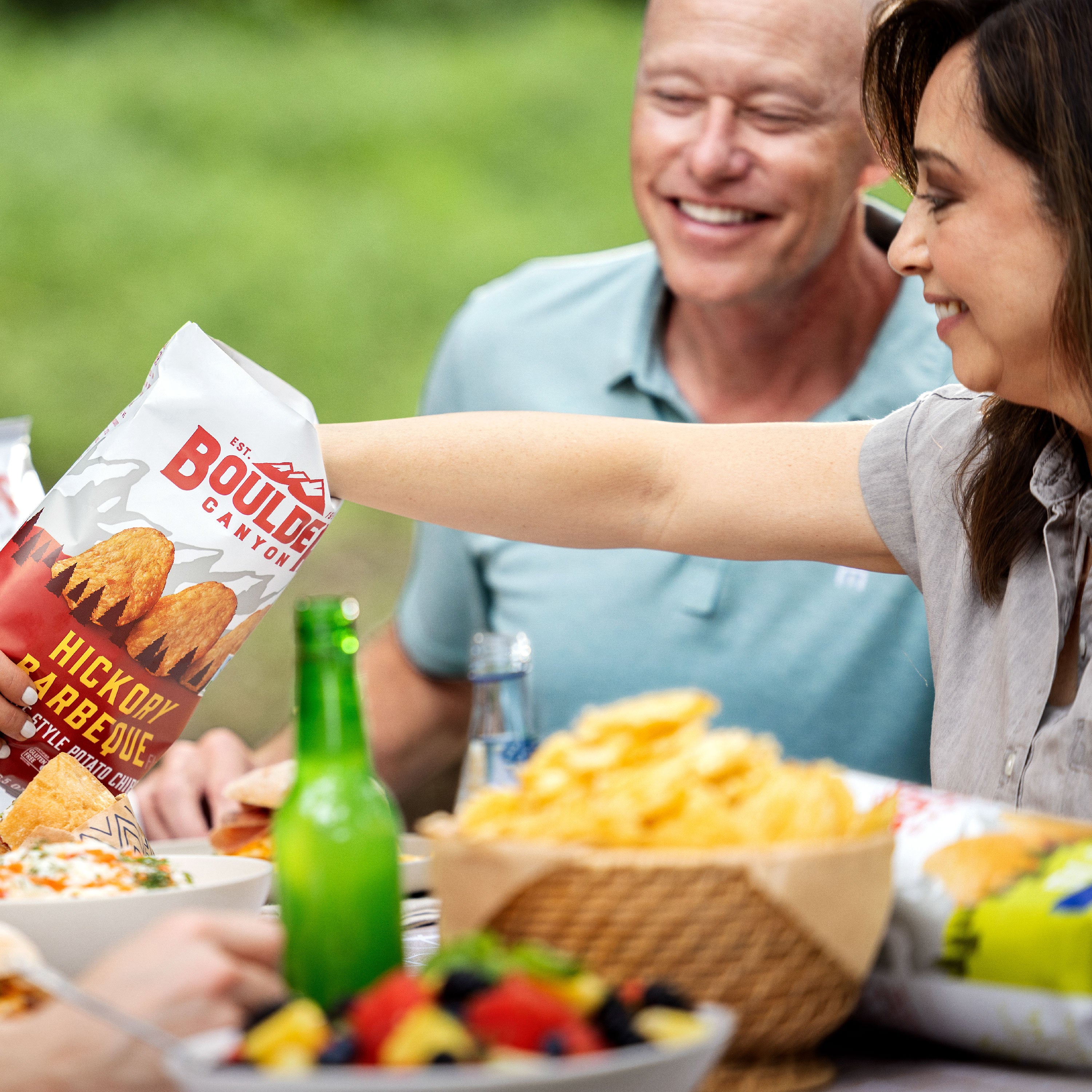Woman reaching for a bag of Boulders Hickory Smoked Barbecue chips at a picnic table with a man in the background.