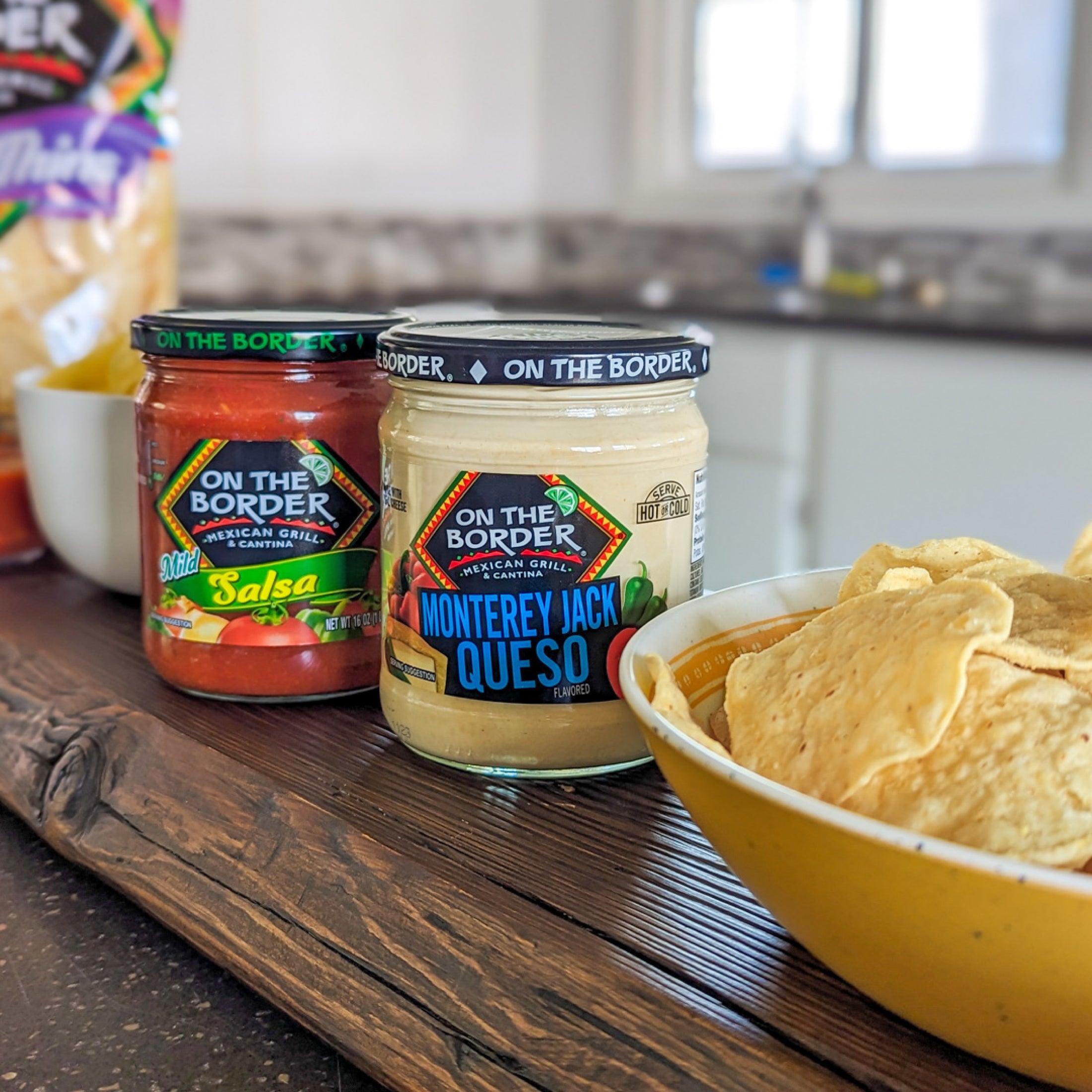 Jars of 'On The Border' salsa and queso with a bowl of chips on a kitchen counter.