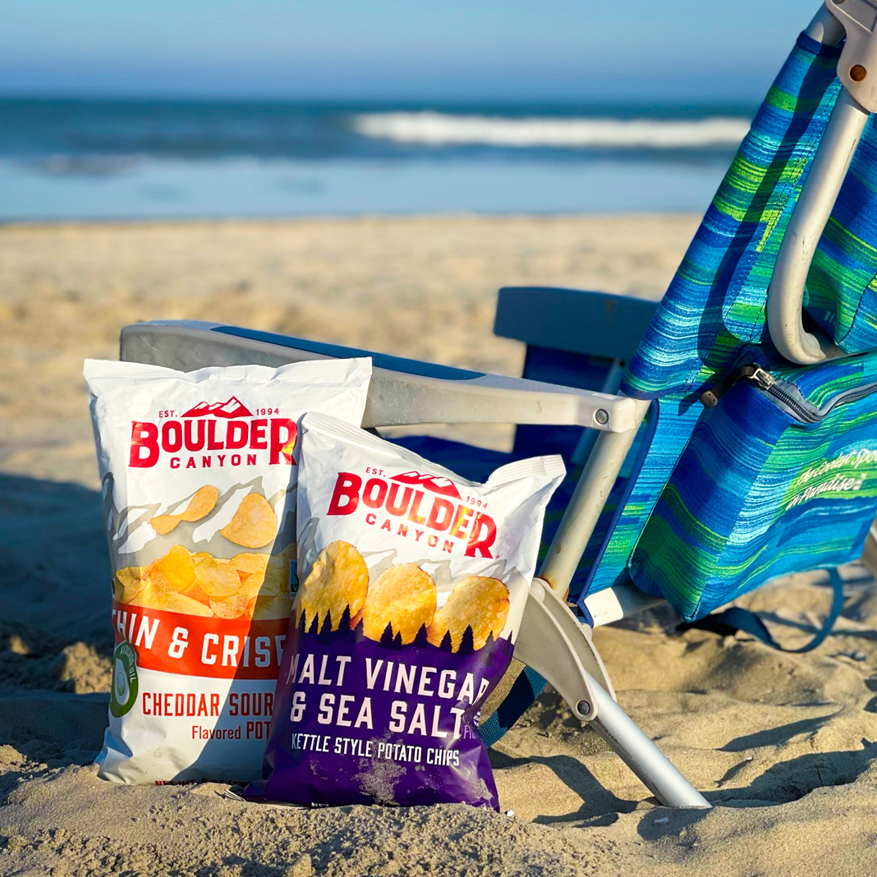 Two bags of Boulder Canyon potato chips on a beach with ocean view