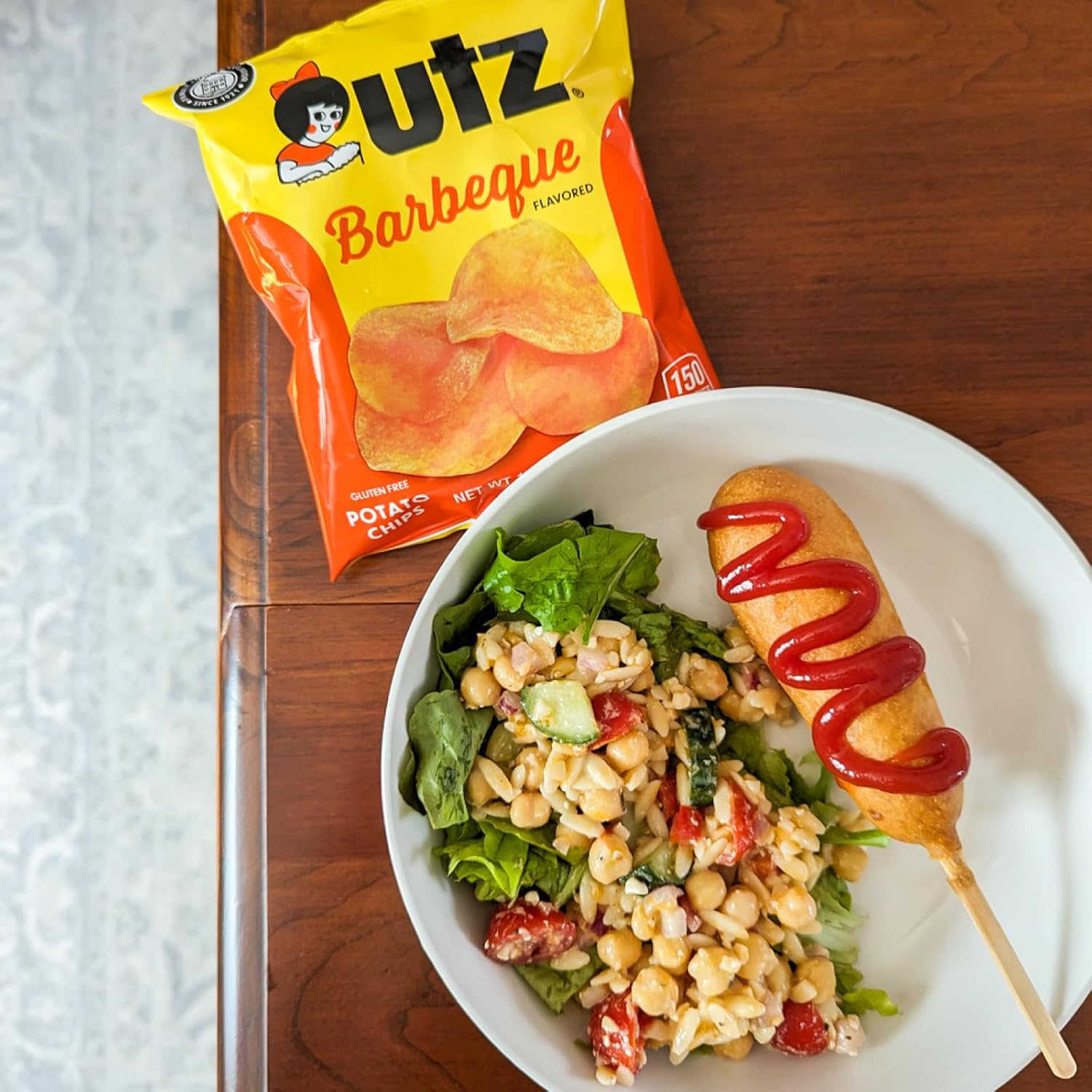 Plate of salad with corn dog and ketchup, next to a bag of Outz Barbeque potato chips on a wooden surface.