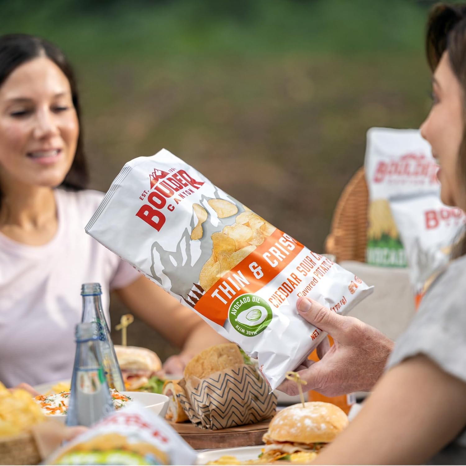 A person offering a bag of Boulder Canyon Thin & Crispy potato chips to another person at a table