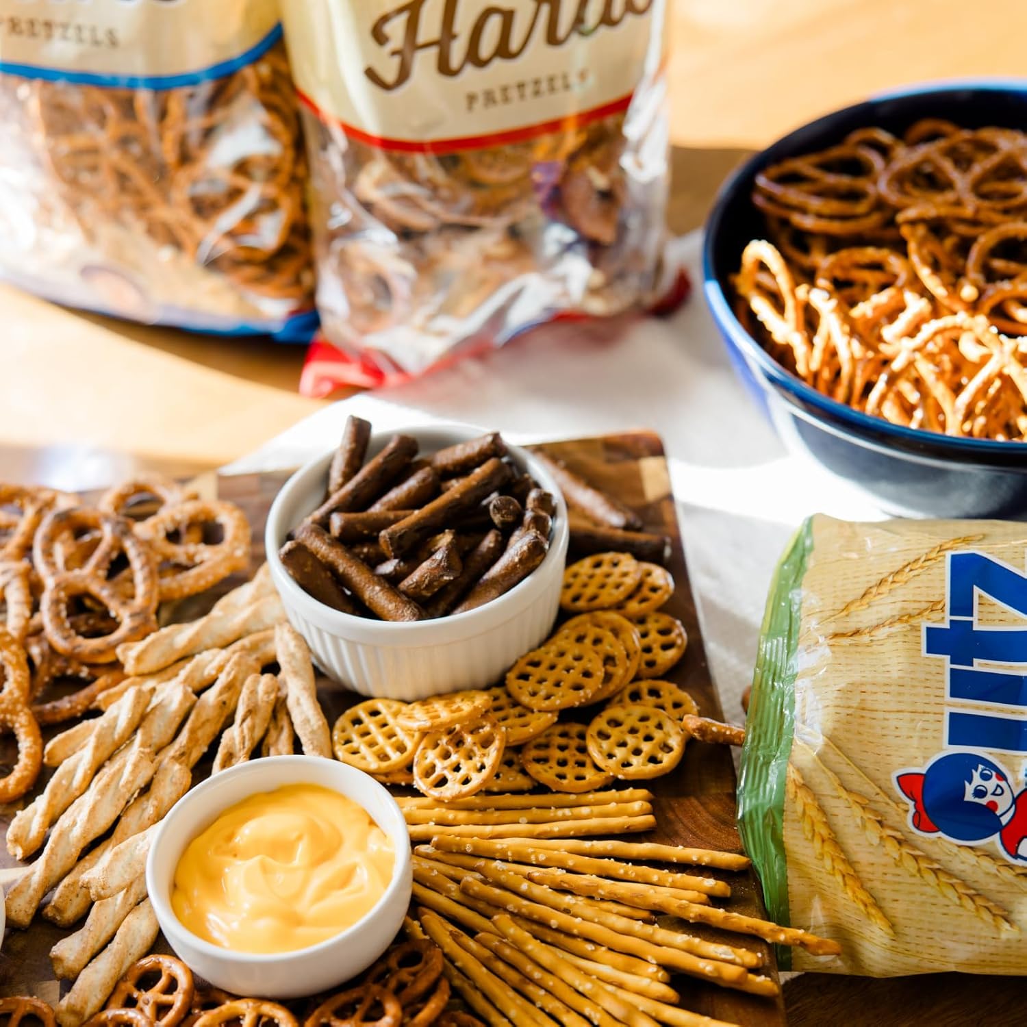 Assorted pretzel snacks with dipping sauce on a wooden board, surrounded by open bags of pretzels.