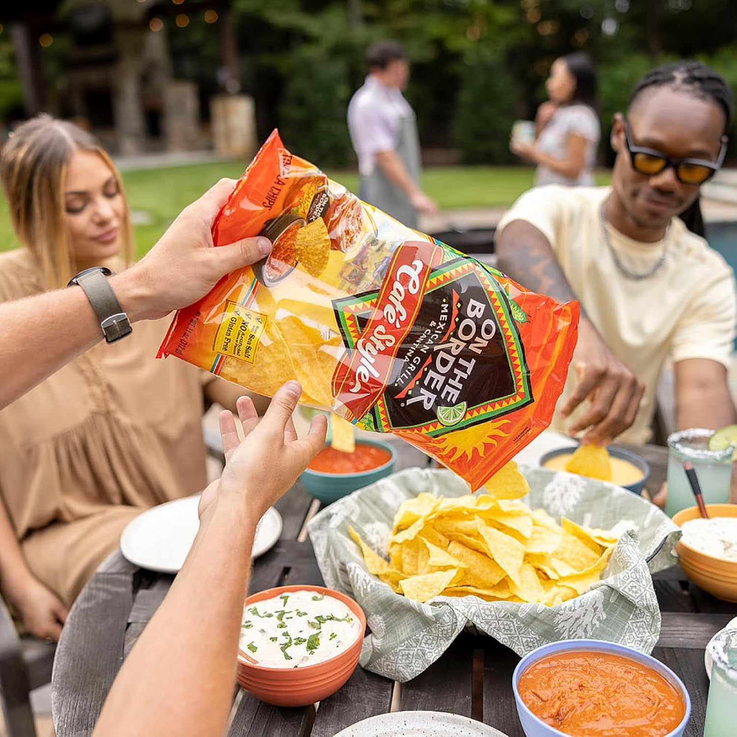 People at a outdoor gathering with a bag of tortilla chips and bowls of salsa.