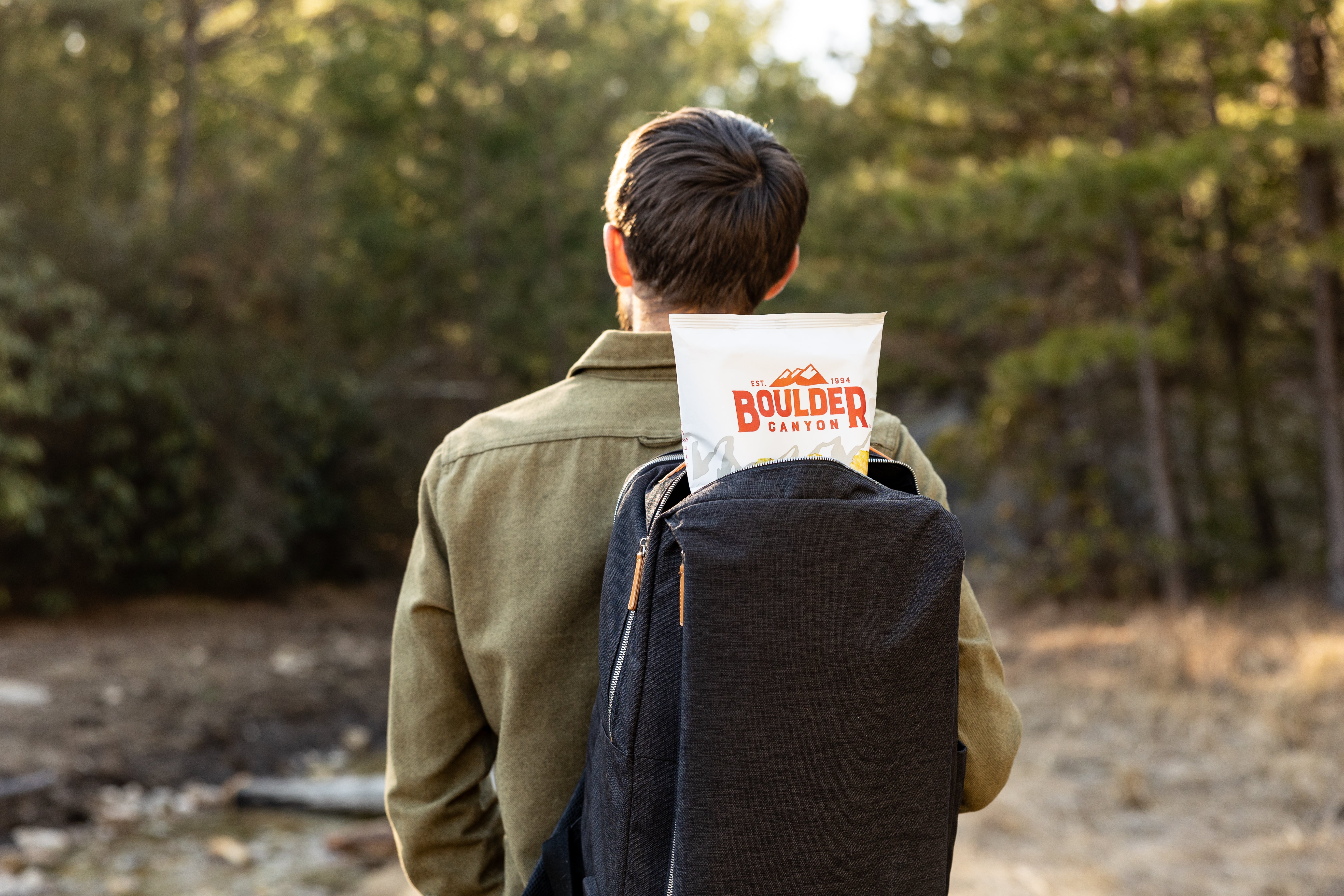 Person with a backpack and Boulder Canyon paper bag in a forest setting