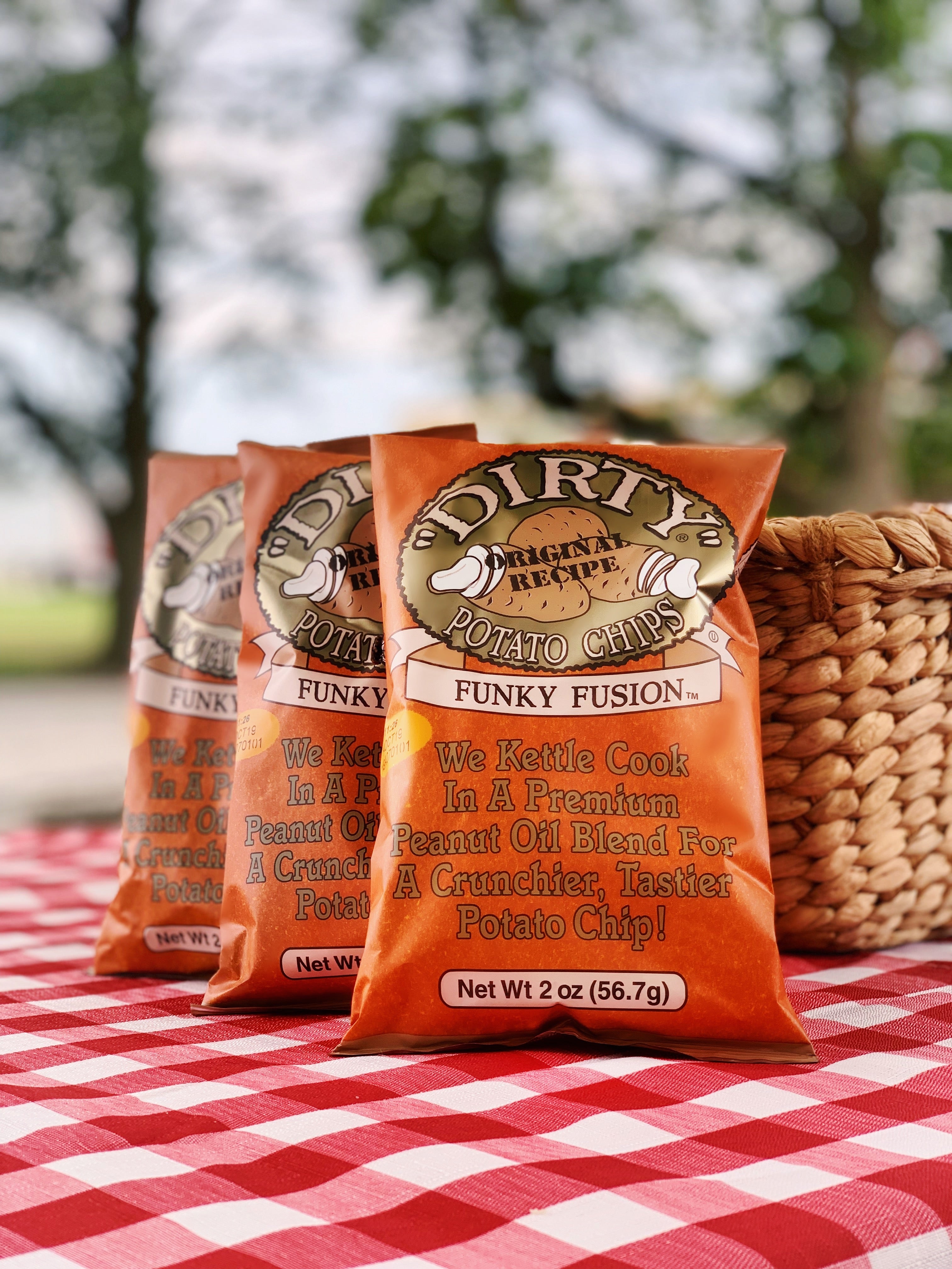 Three bags of Dirty Potato Chips on a red and white checkered tablecloth with a blurred outdoor background.