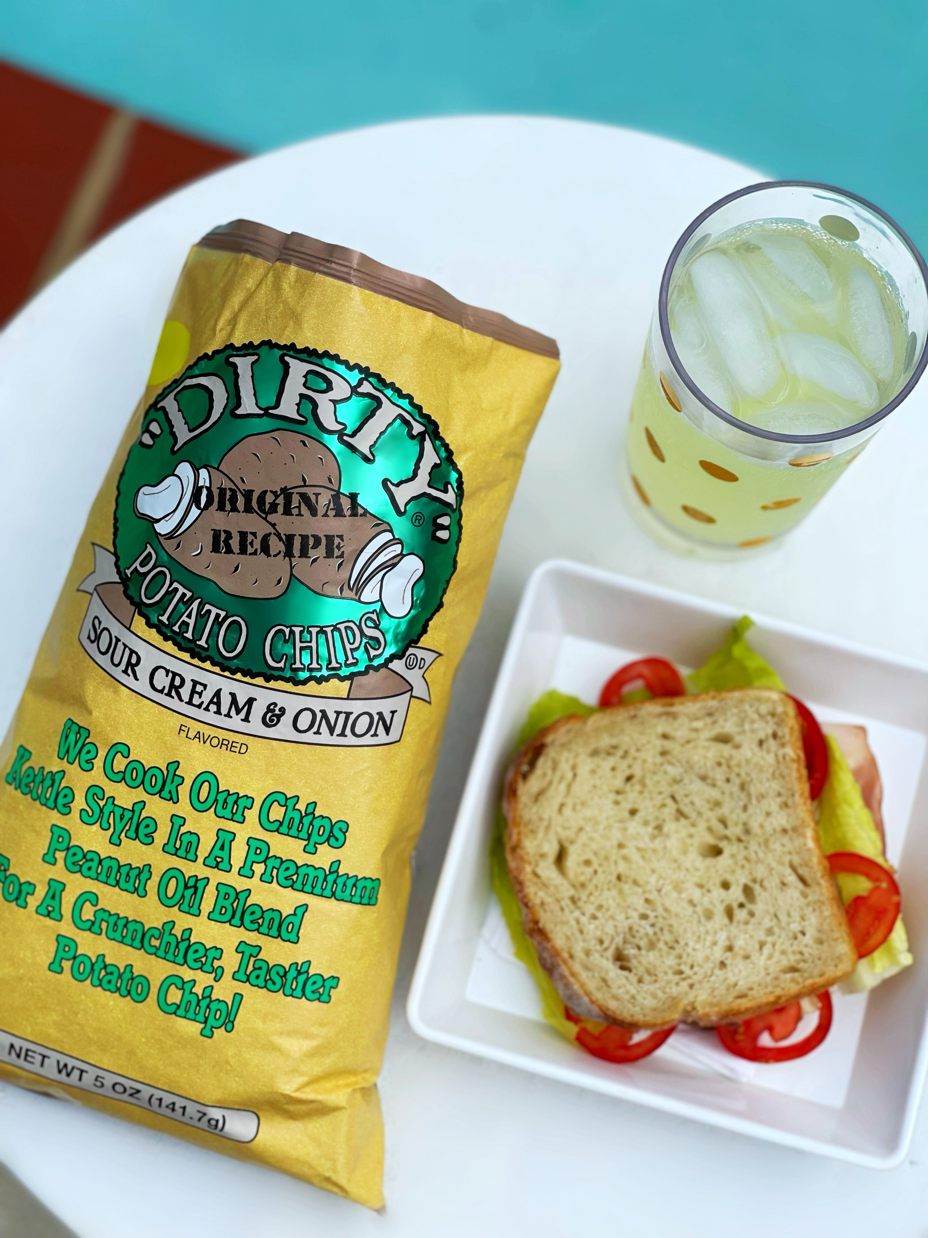 Yellow bag of 'Dirty' potato chips next to a sandwich and a drink on a white plate.