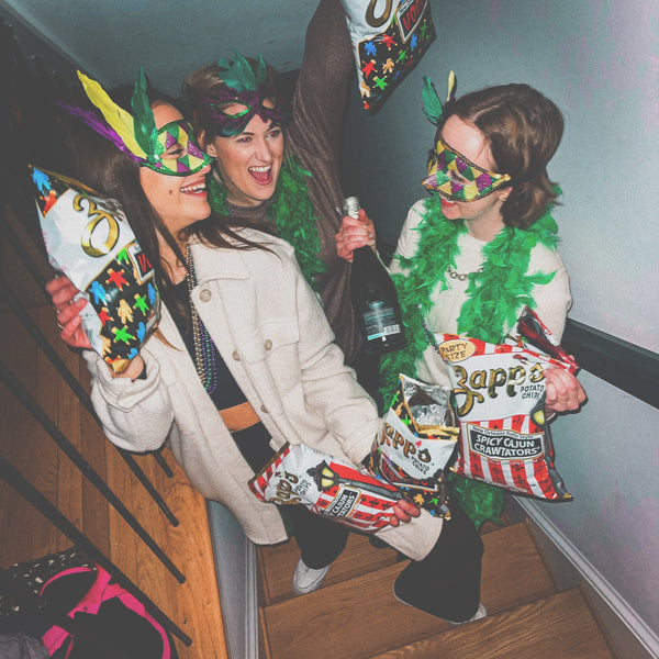 Three people on a staircase wearing festive masks and holding snacks, with a bottle of champagne.
