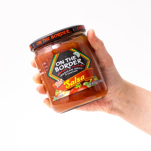 Hand holding a jar of 'On The Border' salsa against a white background