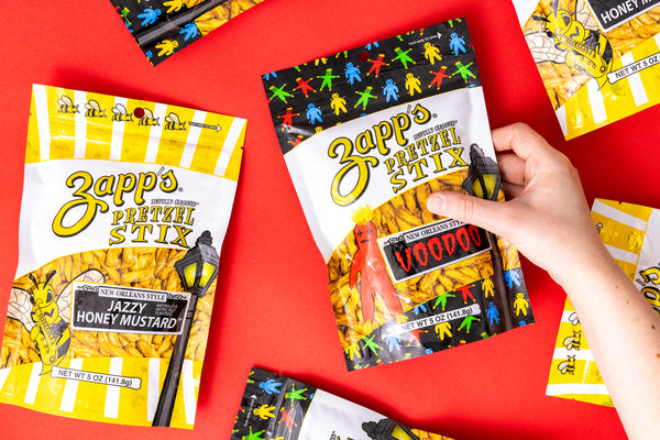 Zapp's Pretzel Stix packages on a red background with a hand holding one package.