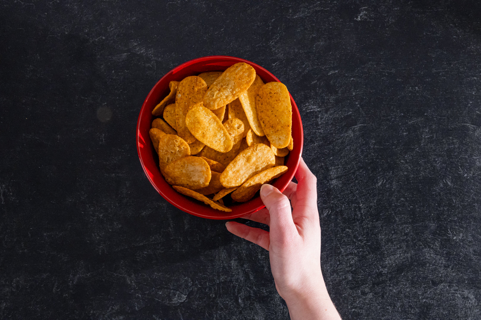 Hand holding a red bowl of potato chips on a dark surface