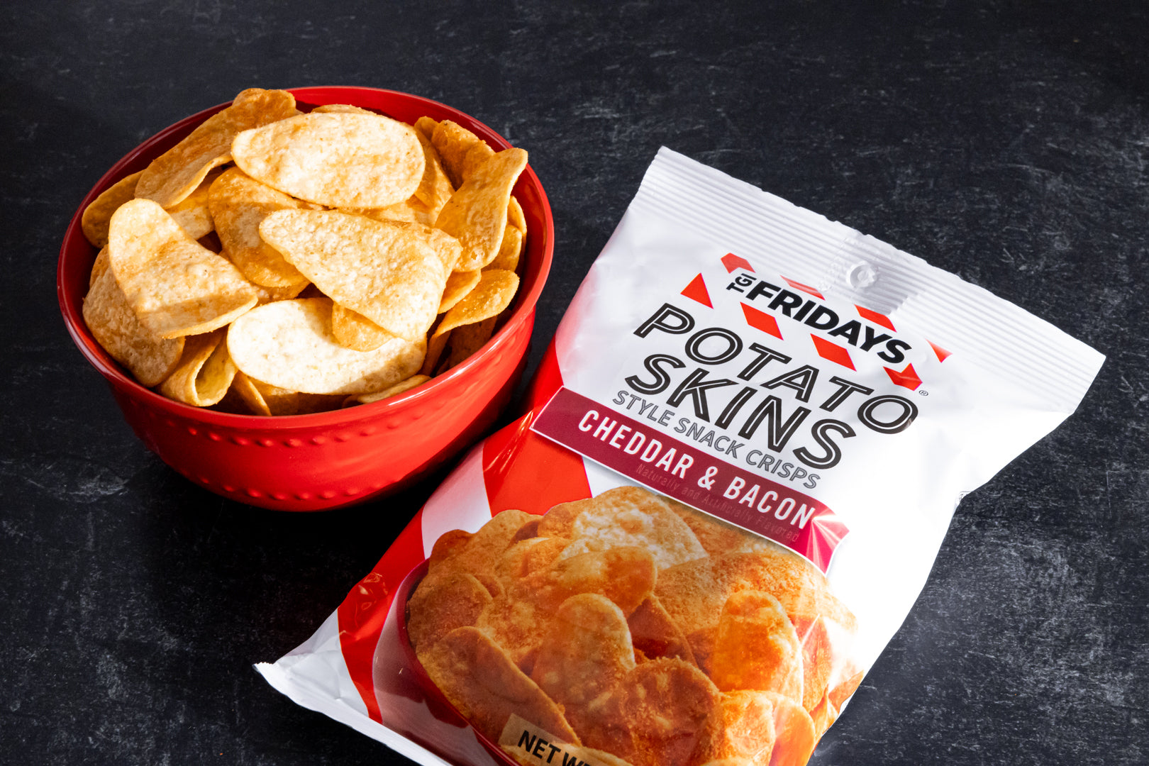 Red bowl of potato skins next to a package of 'Friday's' potato skins on a dark background