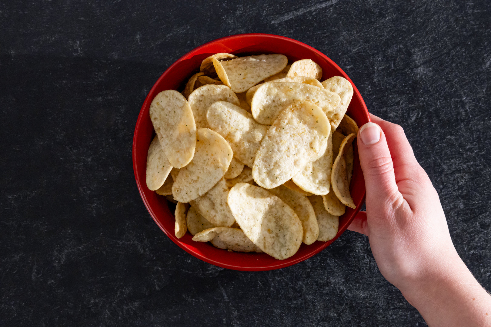 Hand holding a red bowl filled with tortilla chips on a dark surface