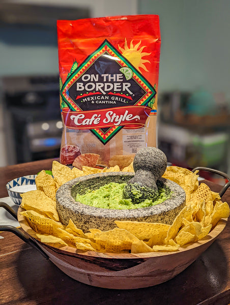 On The Border tortilla chips with a mortar and pestle and guacamole on a wooden board.