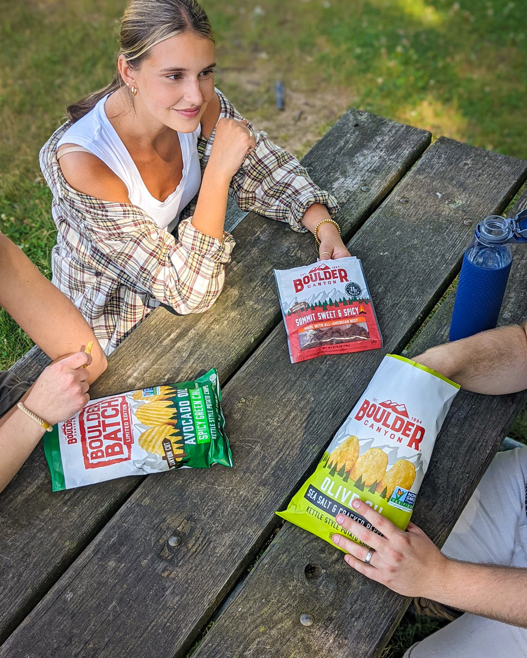 People sitting at a picnic table with Boulder Brand chips and a water bottle.