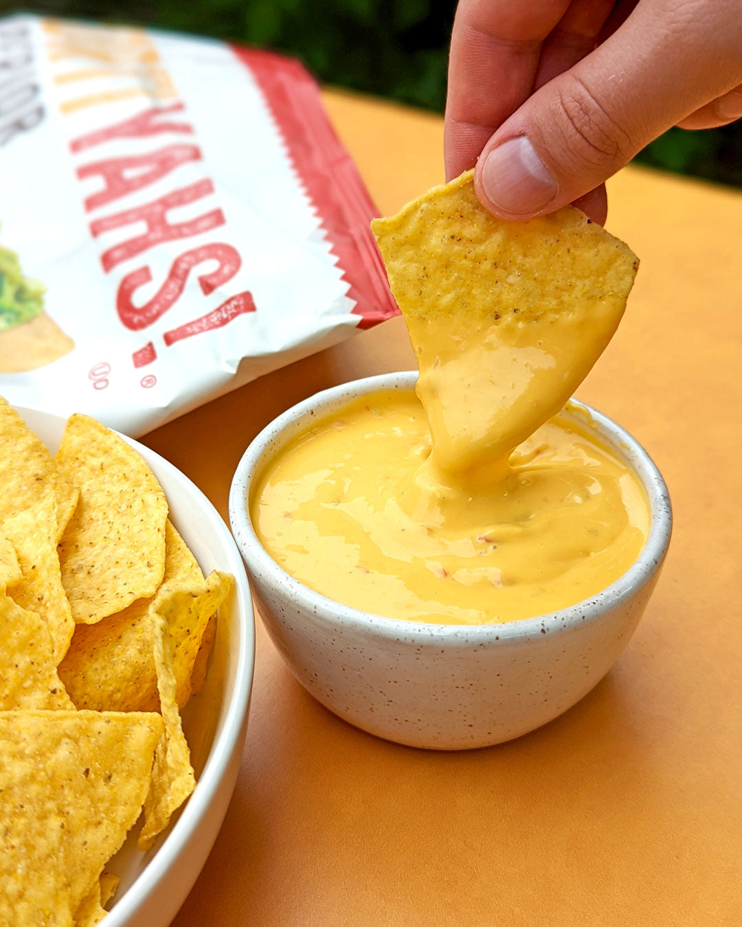 Person dipping a tortilla chip into a bowl of cheese sauce with 'Yahs!' packaging in the background.