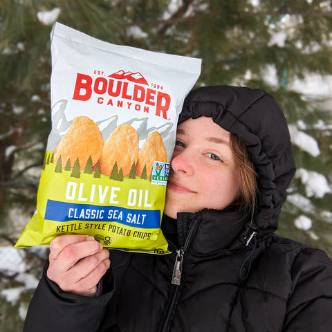 Person holding a Boulder Canyon olive oil kettle-style potato chips bag outdoors with snow and trees in the background