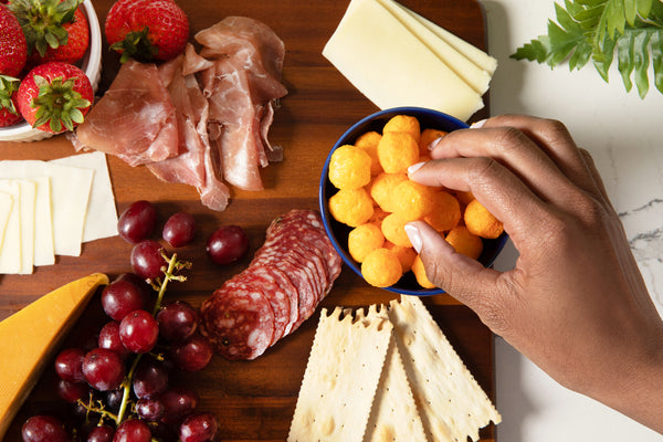 Assorted deli meats, cheeses, fruits, and crackers on a wooden board with a hand reaching for cheese balls.