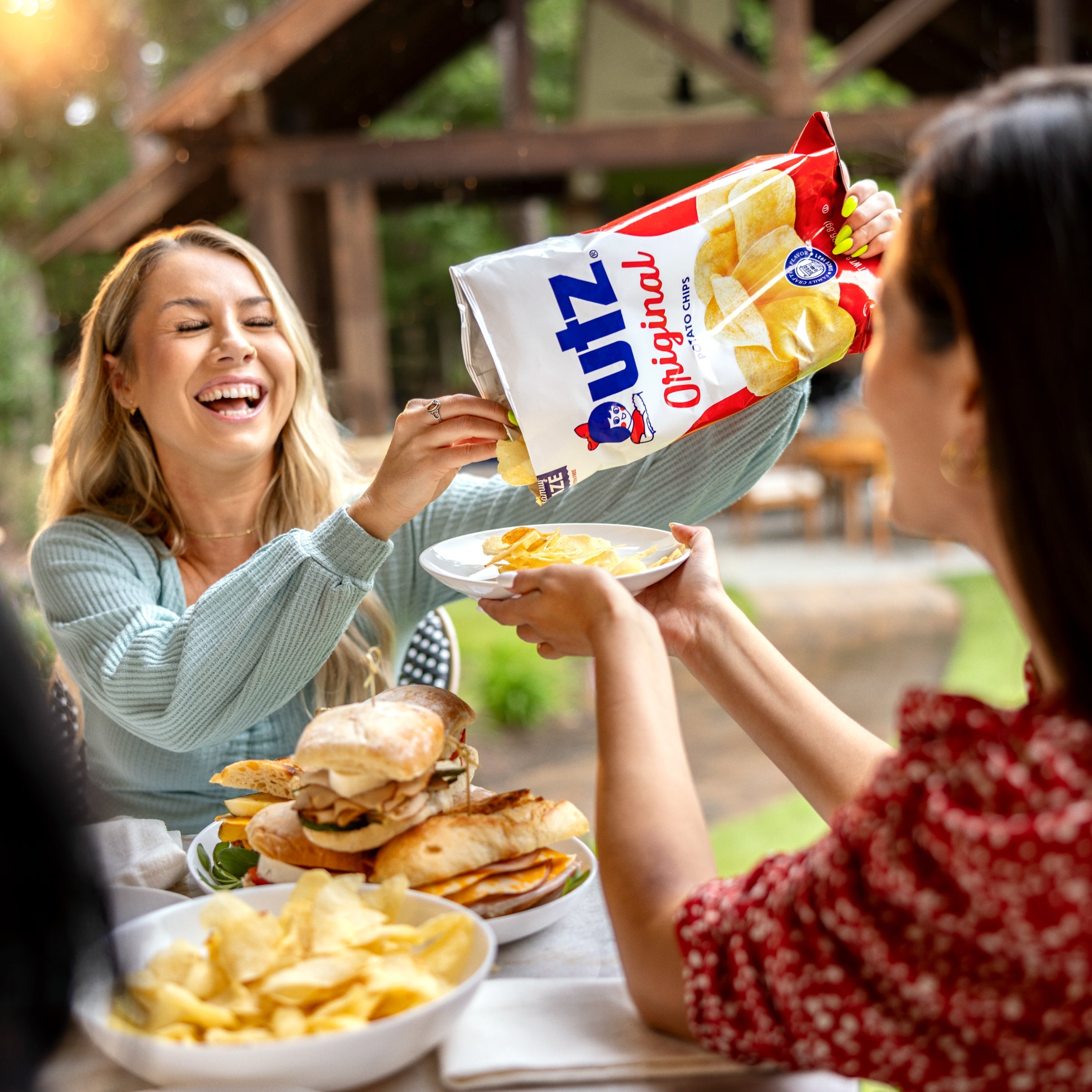A woman smiling as she pours Utz Original Potato Chips from a large bag into a plate held by a friend at an outdoor gathering. The table is set with sandwiches, chips, and other snacks.
