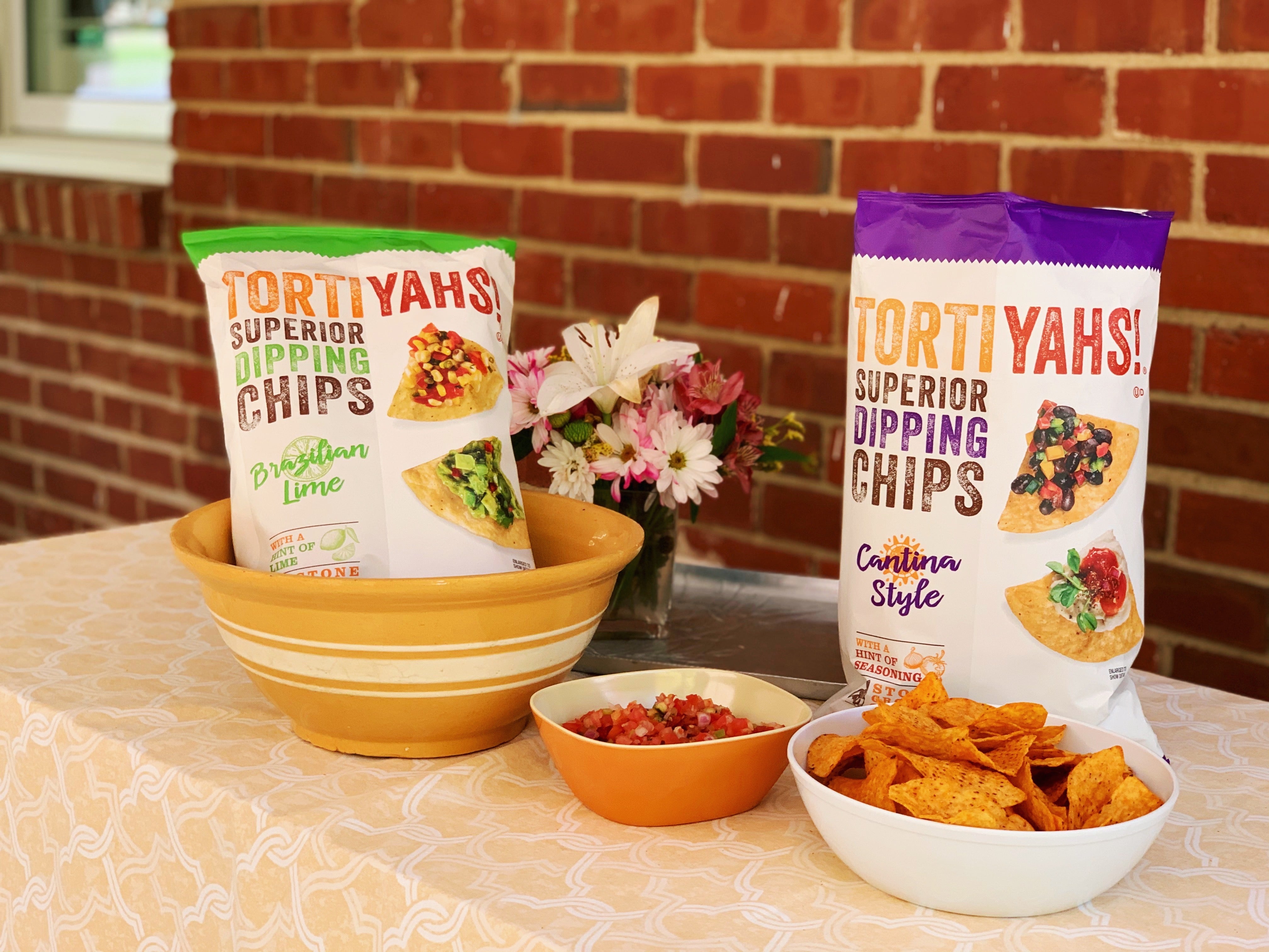 Two bags of Tortiyahs chips on a table with bowls of chips and salsa, against a brick wall.