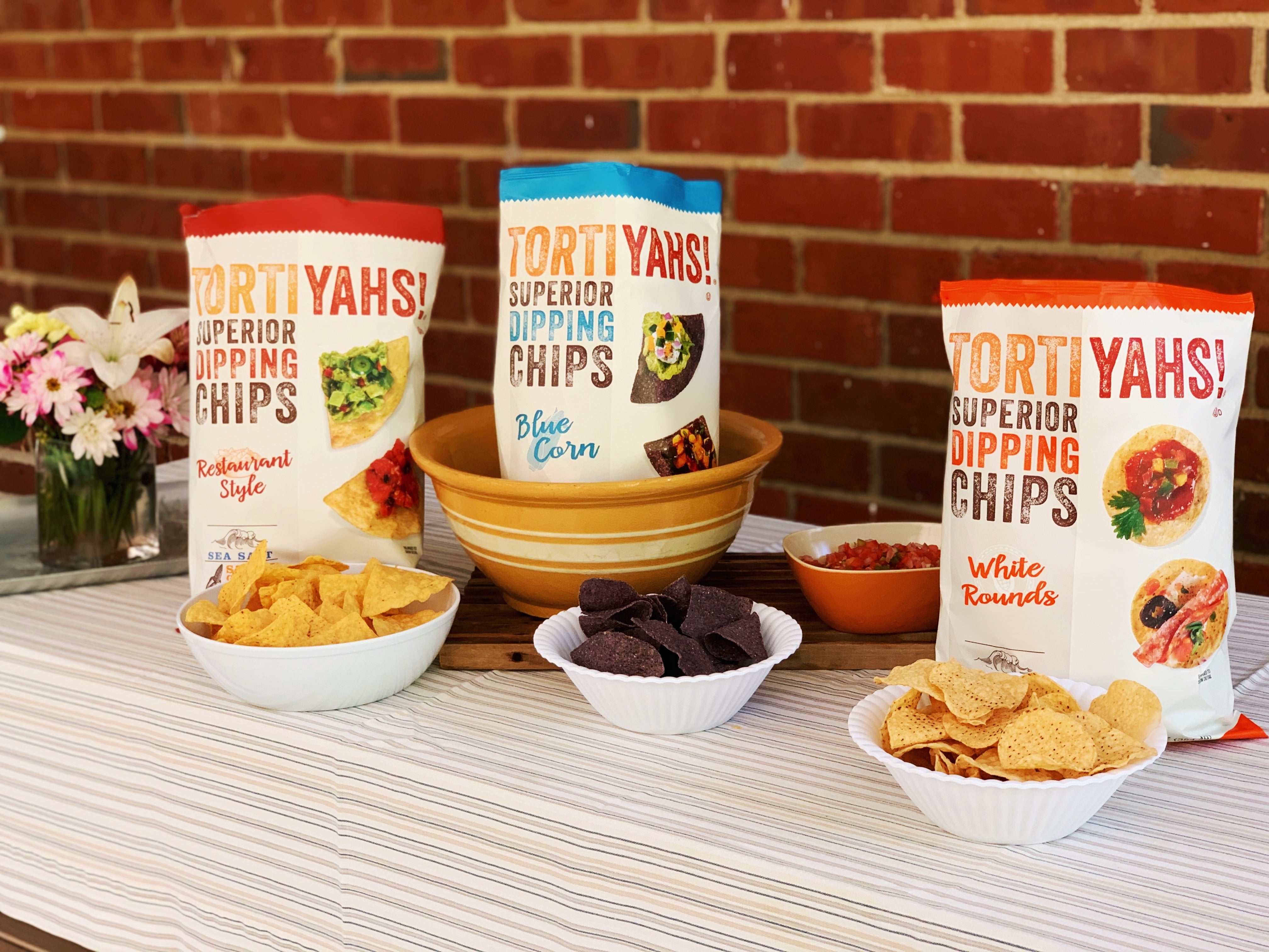 Tortiyahs dipping chips with bowls of chips on a table against a brick wall.