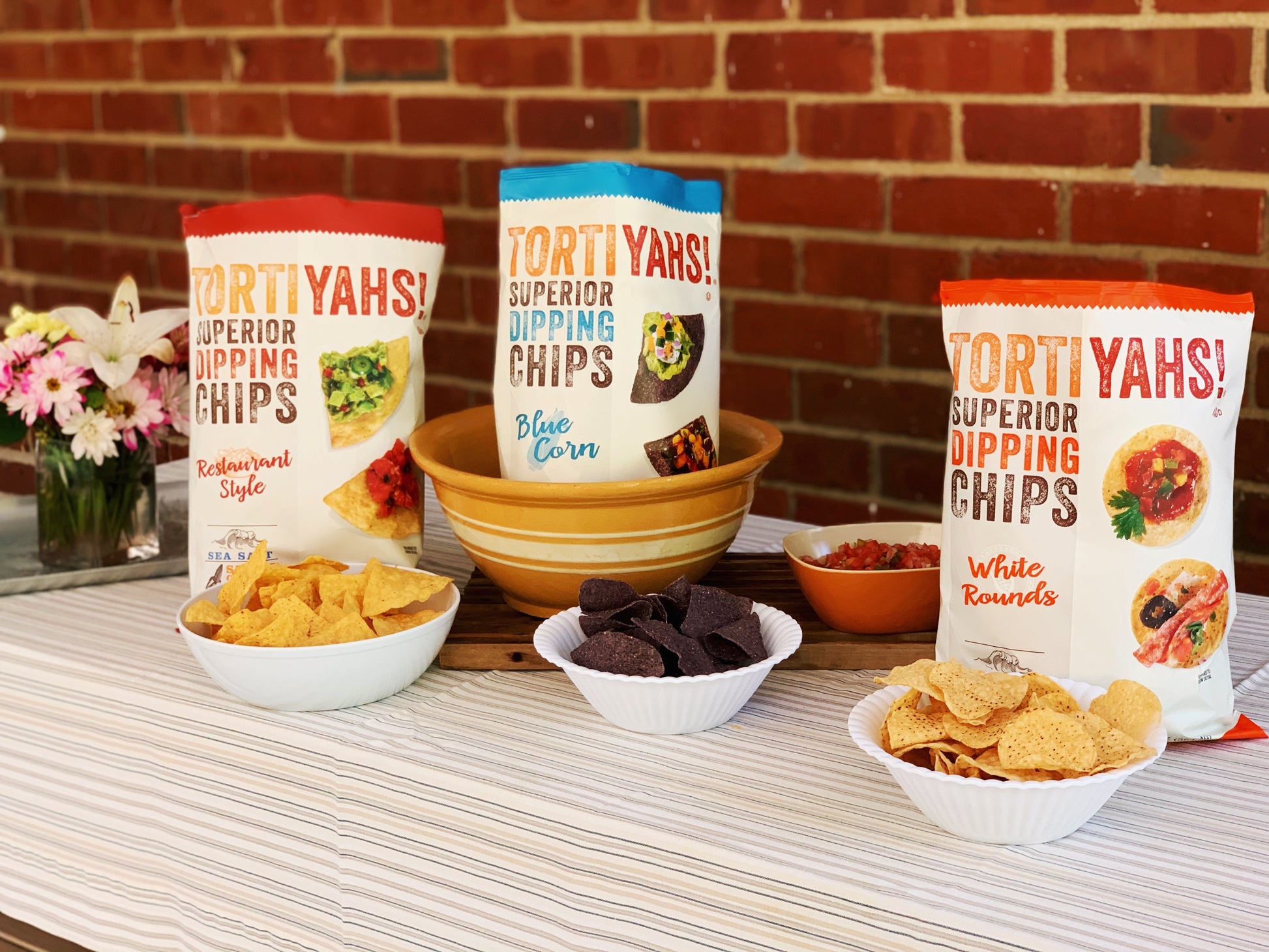Tortiyahs dipping chips with bowls of chips on a table against a brick wall.