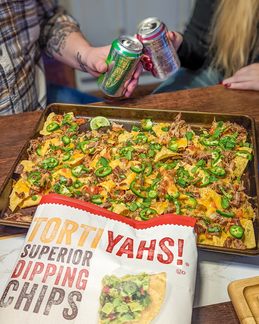Person holding cans of soda with a tray of nachos and a bag of Tortiyahs chips on a table.