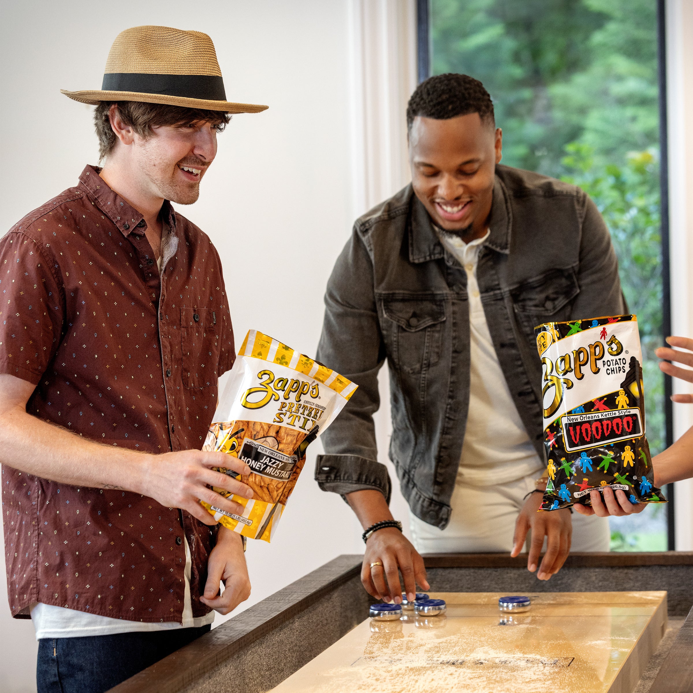 Two men holding Zapp's Pretzel Stix bags (Jazzy Honey Mustard and Voodoo flavors) while playing a game indoors.