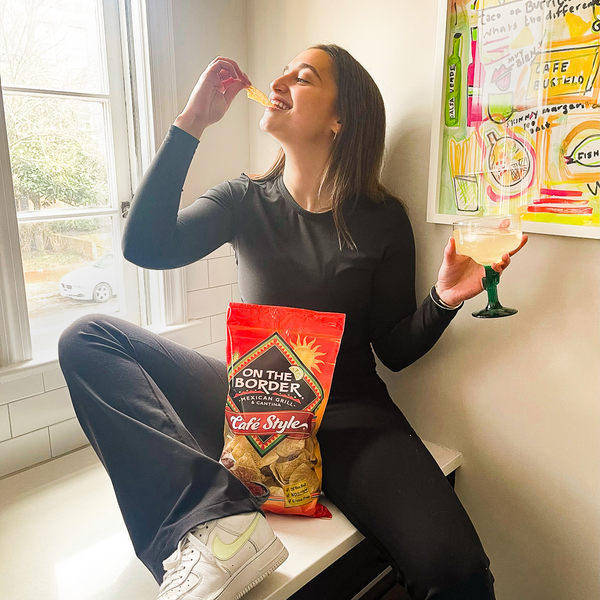 Woman sitting on a windowsill eating chips and drinking a beverage, with 'On the Border' chips visible.