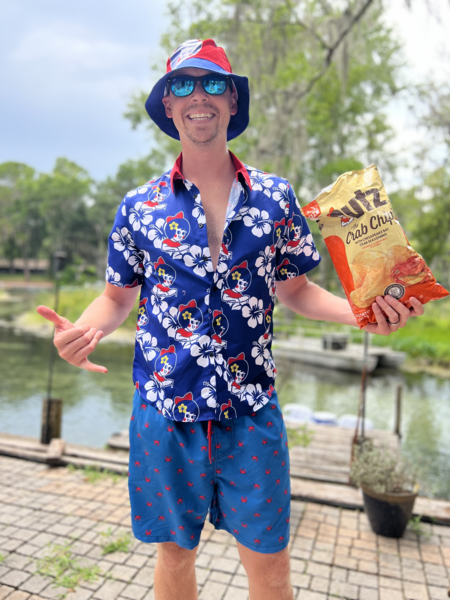 Person wearing a colorful shirt and shorts holding a bag of chips outdoors.